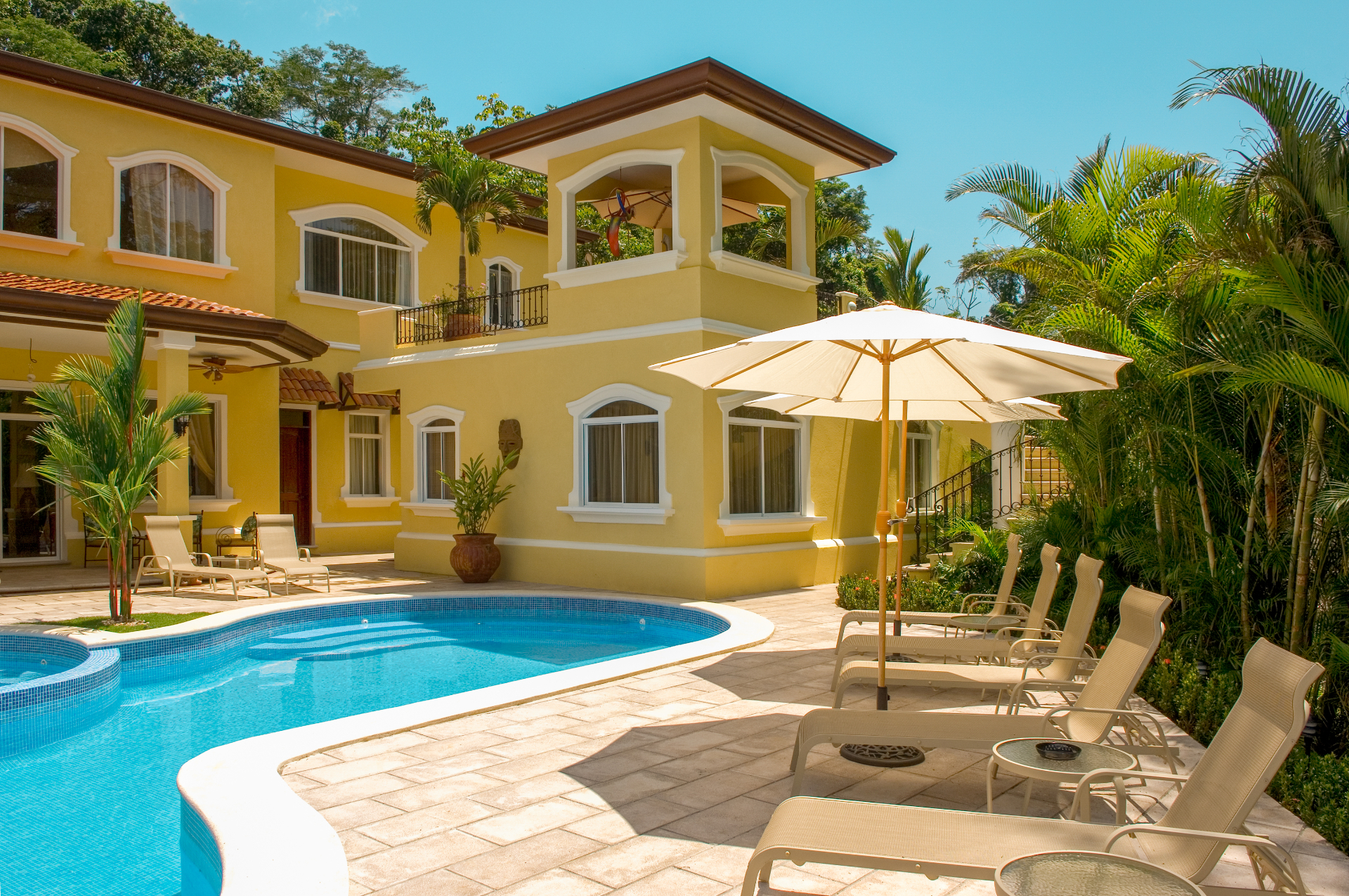 Private pool terrace with sun loungers and umbrella beside a yellow Los Sueños resort villa.