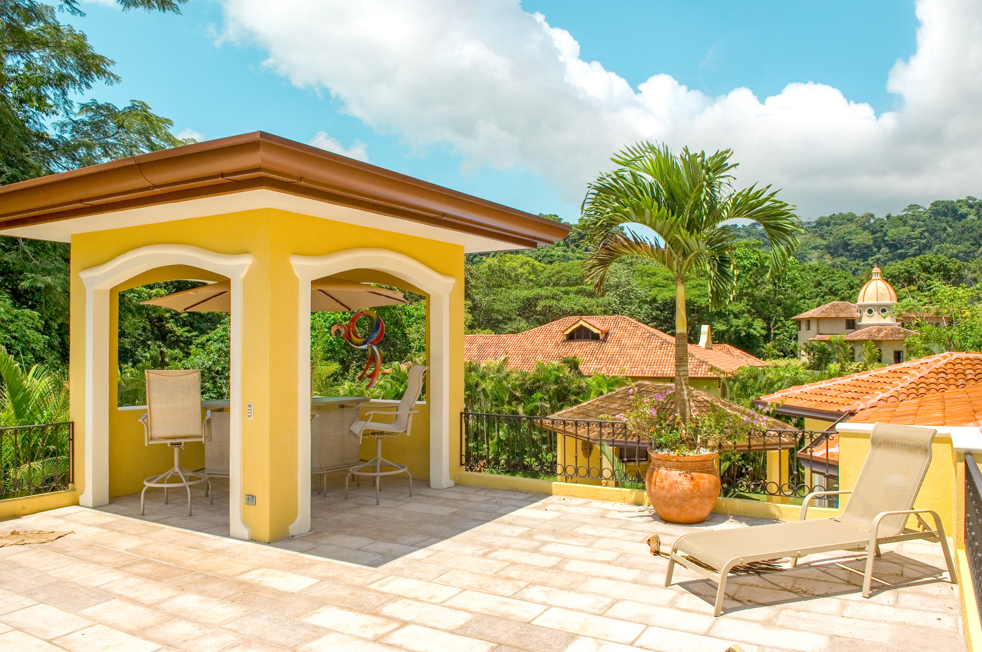 Rooftop terrace with lounge chair and covered seating area overlooking tiled roofs and tropical hills in Los Sueños resort.