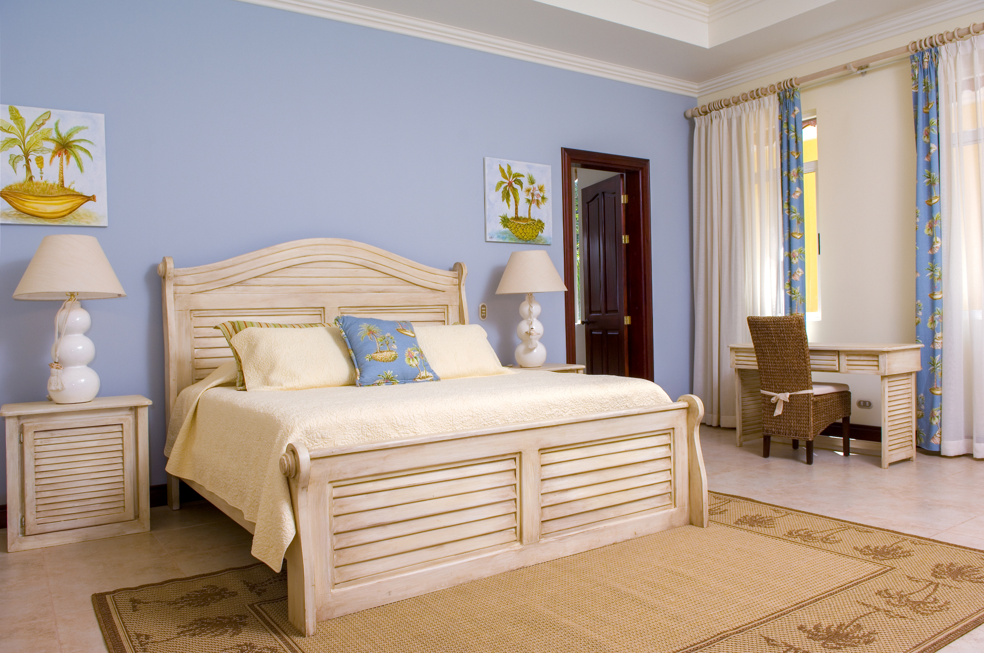 King bedroom with light wood bed, blue accent wall, bedside lamps, desk, and window with curtains inside a Los Sueños villa.