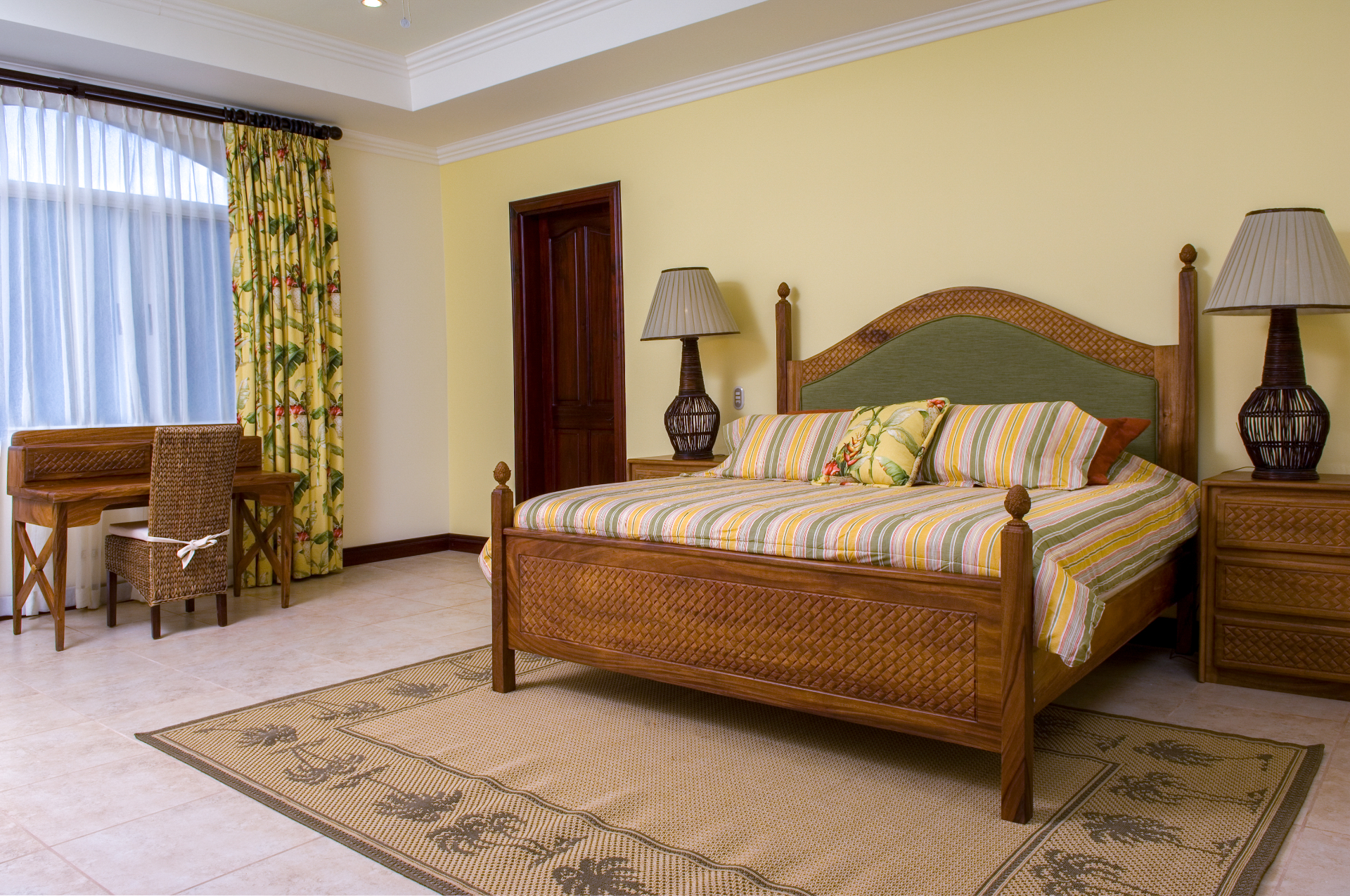 King bedroom with wooden bed frame, bedside lamps, desk, and window with tropical curtains inside a Los Sueños villa.