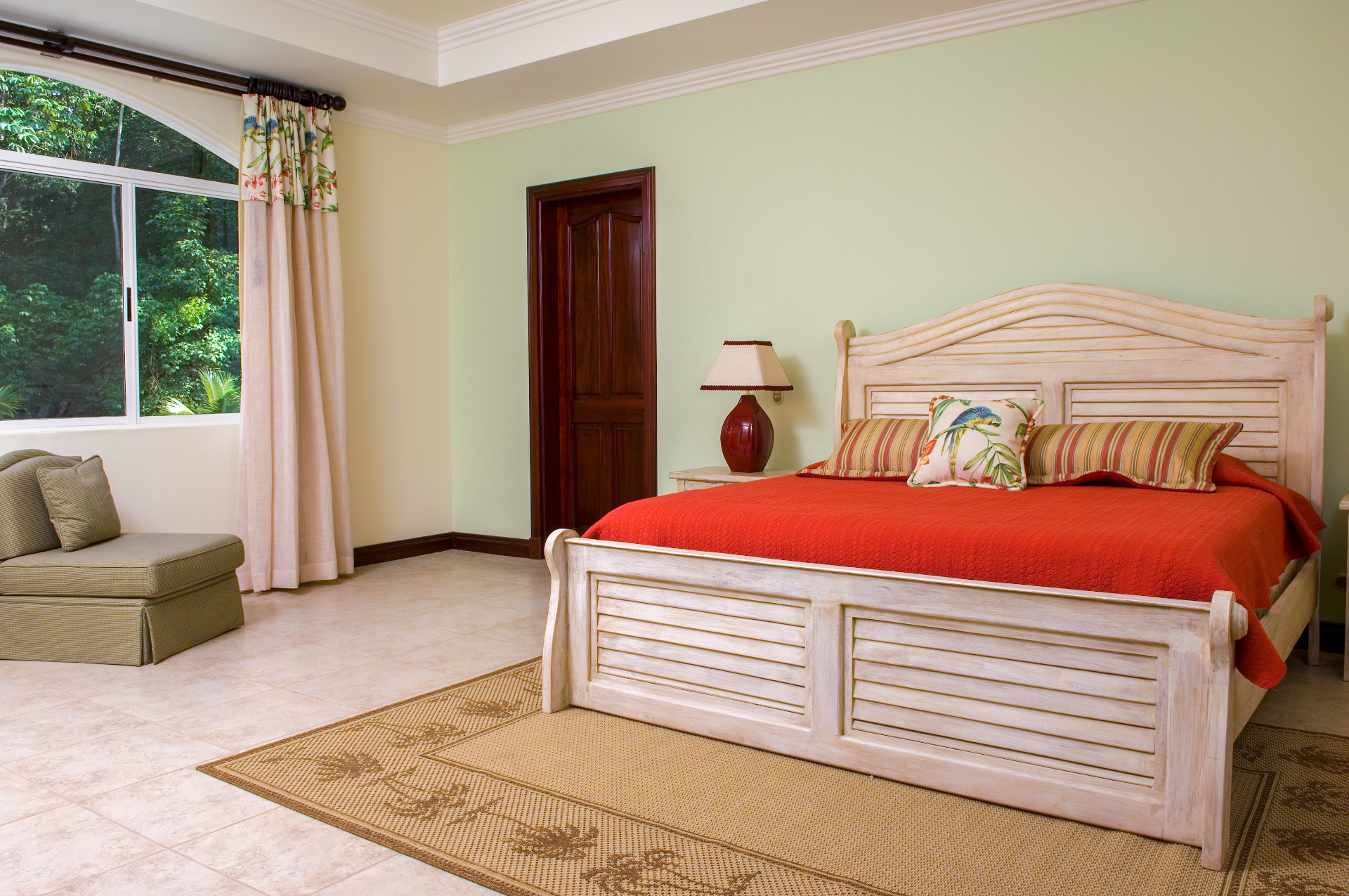 King bedroom with white wood bed frame, red bedding, and large window overlooking tropical greenery in a Los Sueños villa.