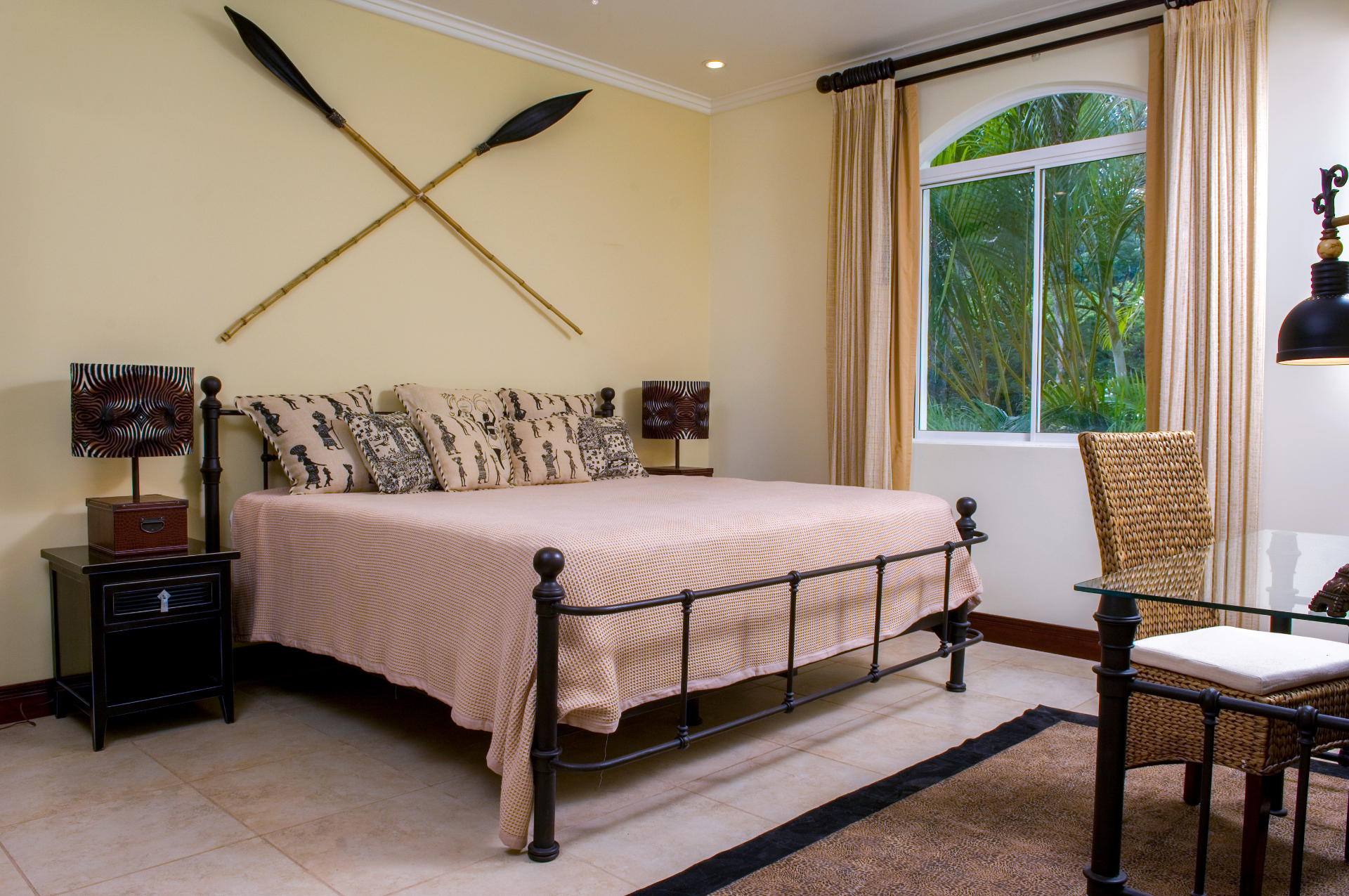 King bedroom with metal bed frame, decorative paddles on the wall, desk, and window overlooking tropical garden at a Los Sueños villa.