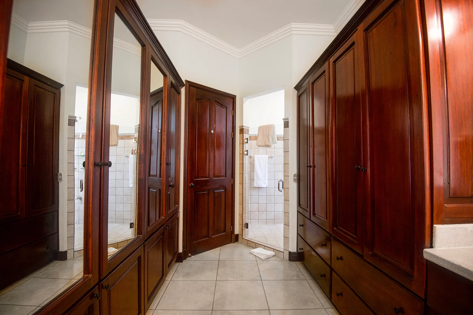 Walk-in closet with built-in wooden wardrobes and mirrors leading to an ensuite bathroom with tiled shower in a Los Sueños villa.