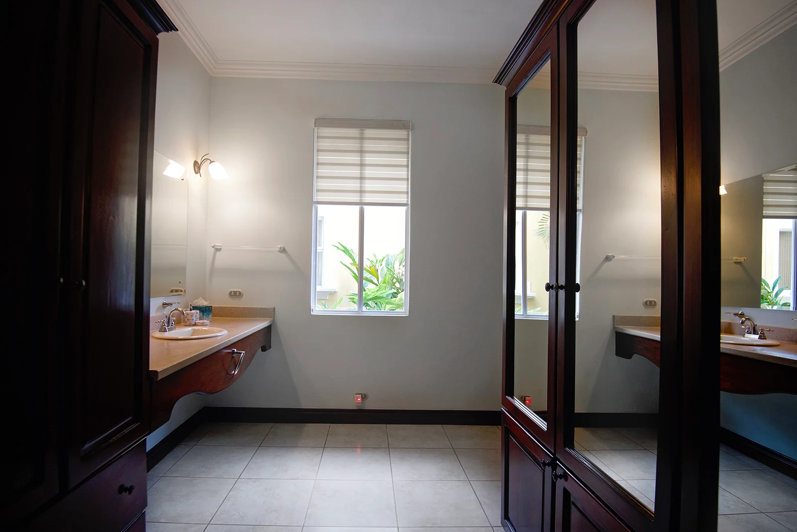 Ensuite bathroom with sink vanity, mirror, and window overlooking tropical plants inside a Los Sueños villa.