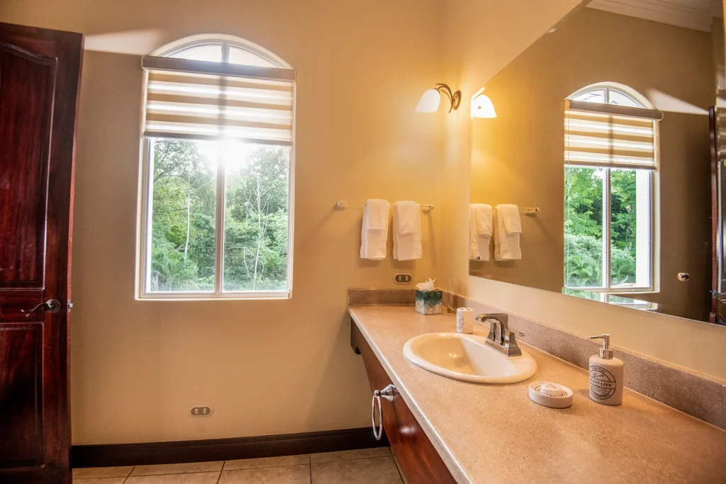 A bright bathroom vanity with large mirror and window overlooking tropical greenery.