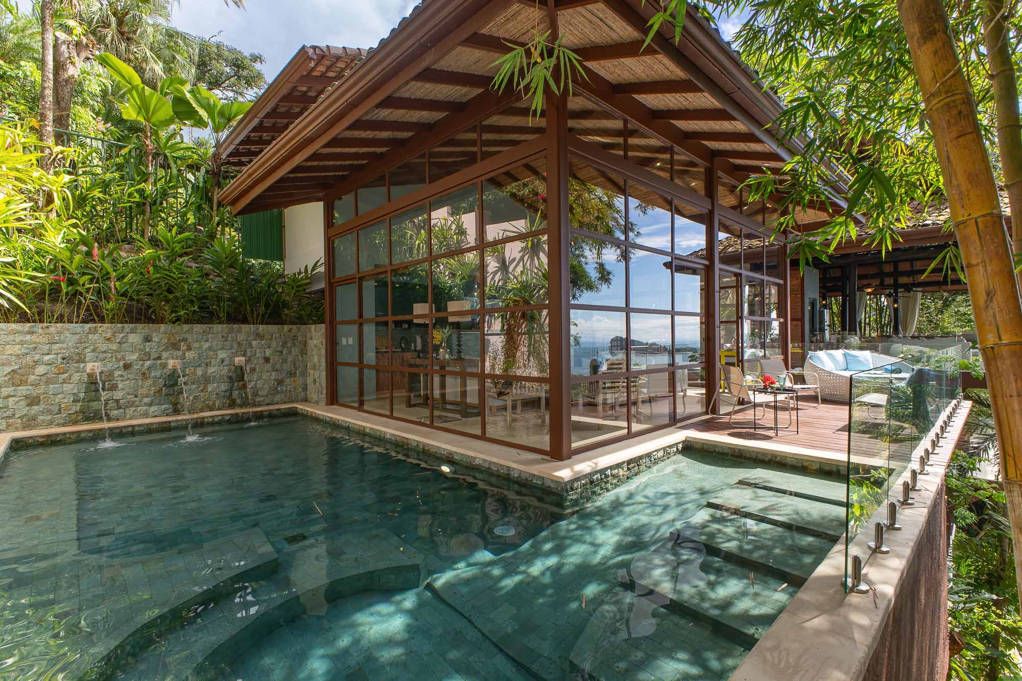 Poolside view of the newly built wing with glass walls, stone details, wooden structure, pool water, and surrounding jungle at a villa in Manuel Antonio, Costa Rica.