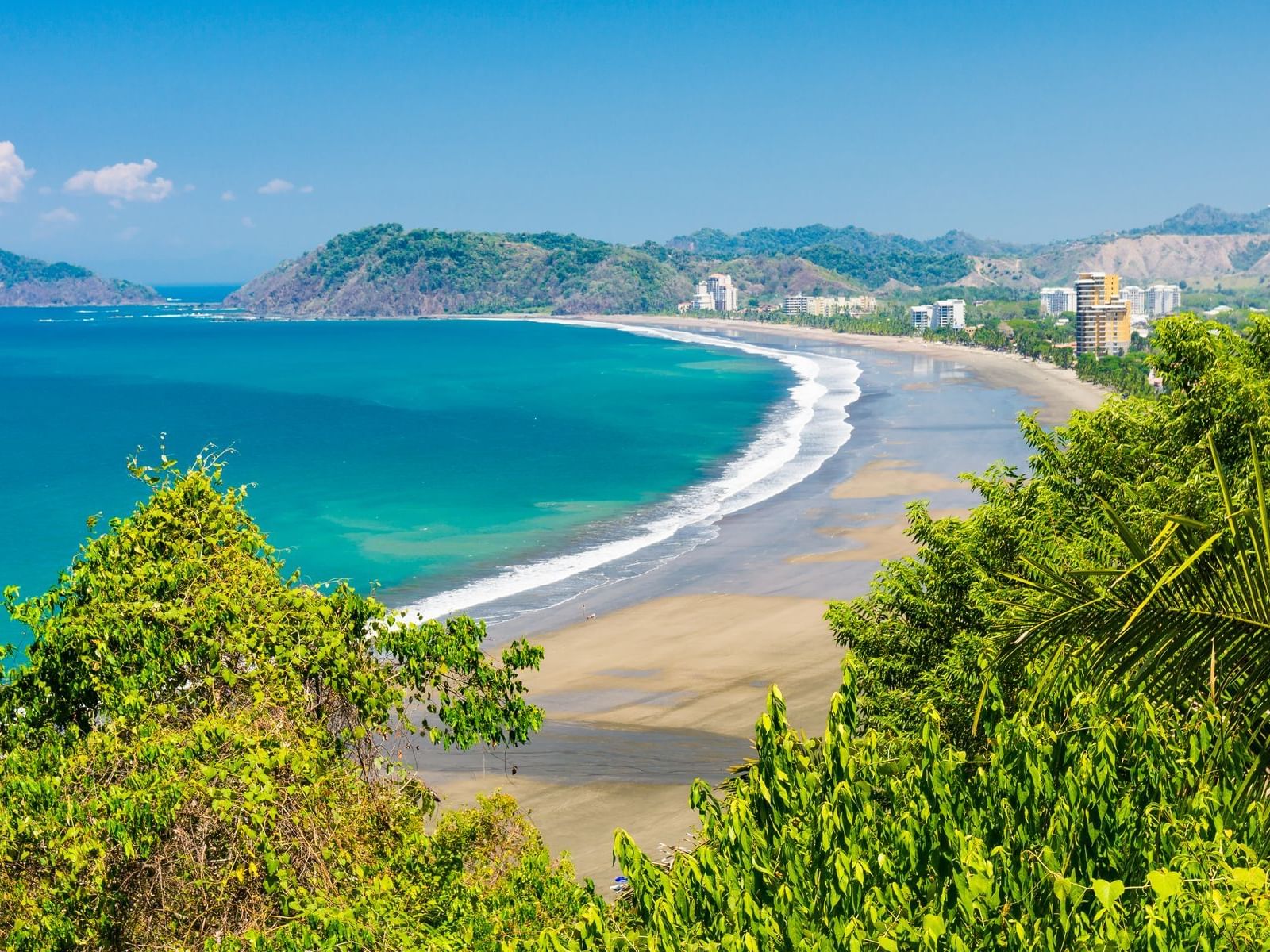 Aerial view of Jacó Beach coastline with ocean waves, sandy shore, and surrounding hills.