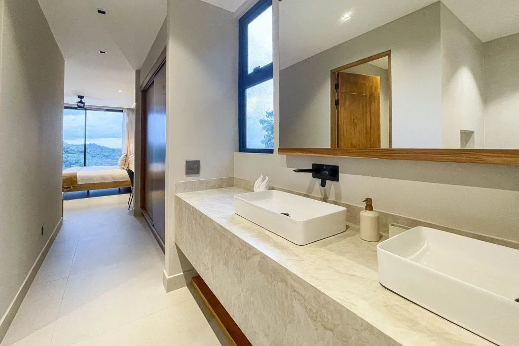 This modern ensuite bathroom has a large closet and double sinks on a beautiful stone counter.