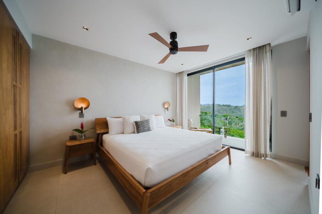 A calm, light-filled bedroom opening to lush jungle and ocean views just beyond the glass.